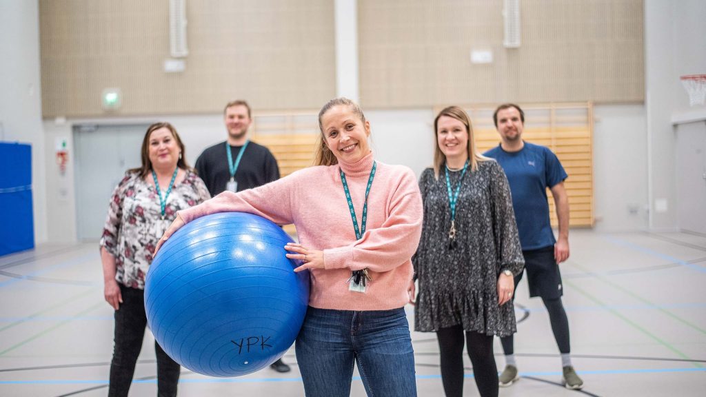 Viisi opettajaa avainnauhat kaulassaan jumppasalissa. Etummaisella jumppapallo kainalossaan.